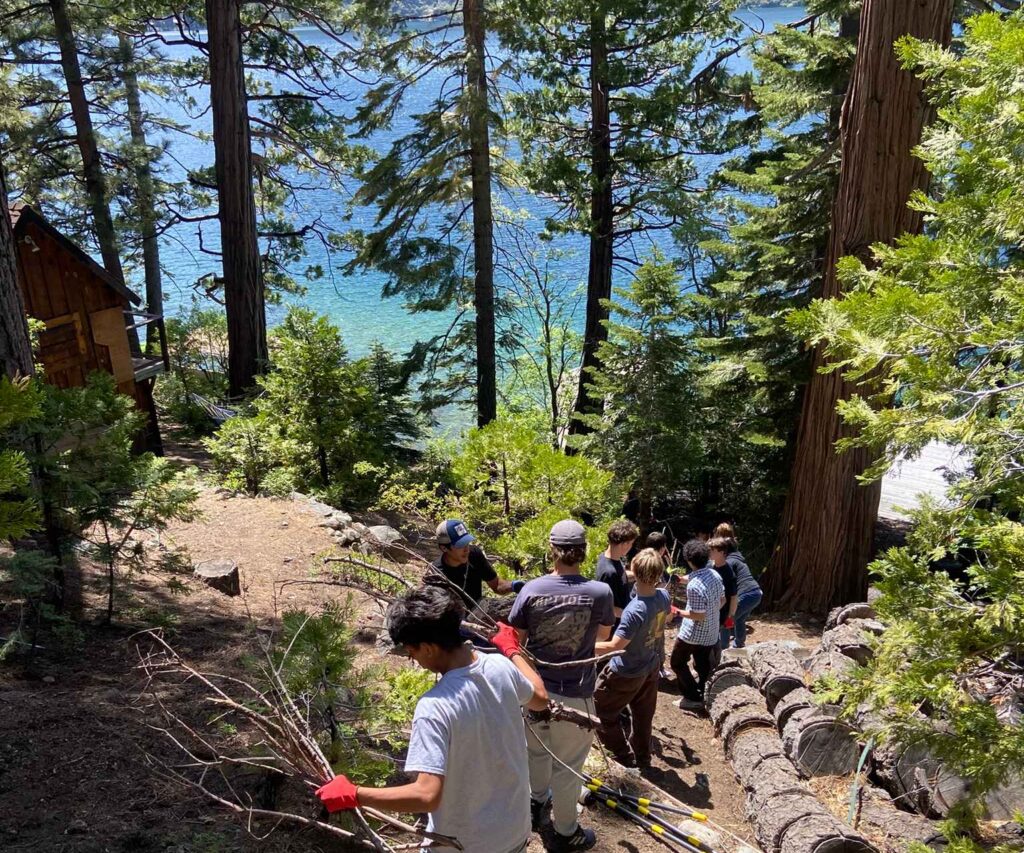 Line of high school students passing dry forest fuels up a slope to reduce wildfire risk. Features the bright teal/blue Fallen Leaf Lake behind tall pines near the water.
